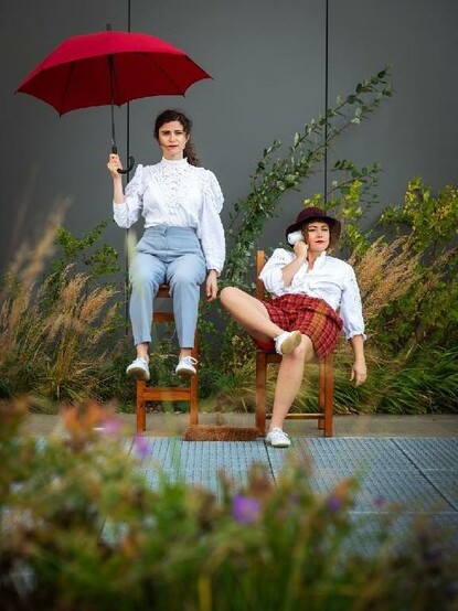 zwei Frauen auf einem Stuhl mit Regenschirm und Föhn in der Hand.