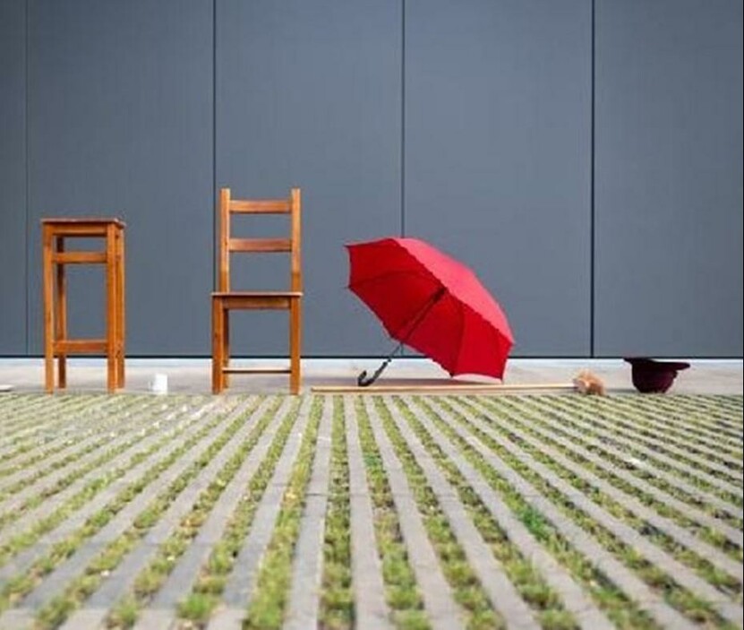 Hocker, Stuhl, Regenschirm, Besen, Kaffeetasse und Hut im Freien vor einer grauen Außenwand